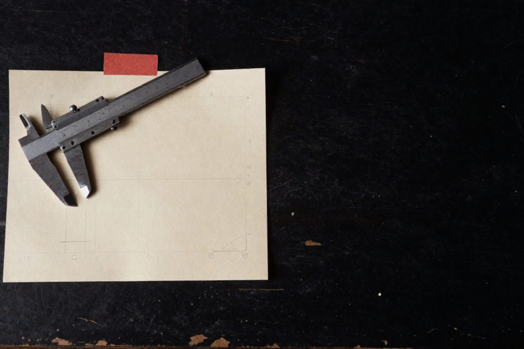 Vintage calipers on a cream draughting template with pencil lines, small clay-red paper tab, dark oak workbench.