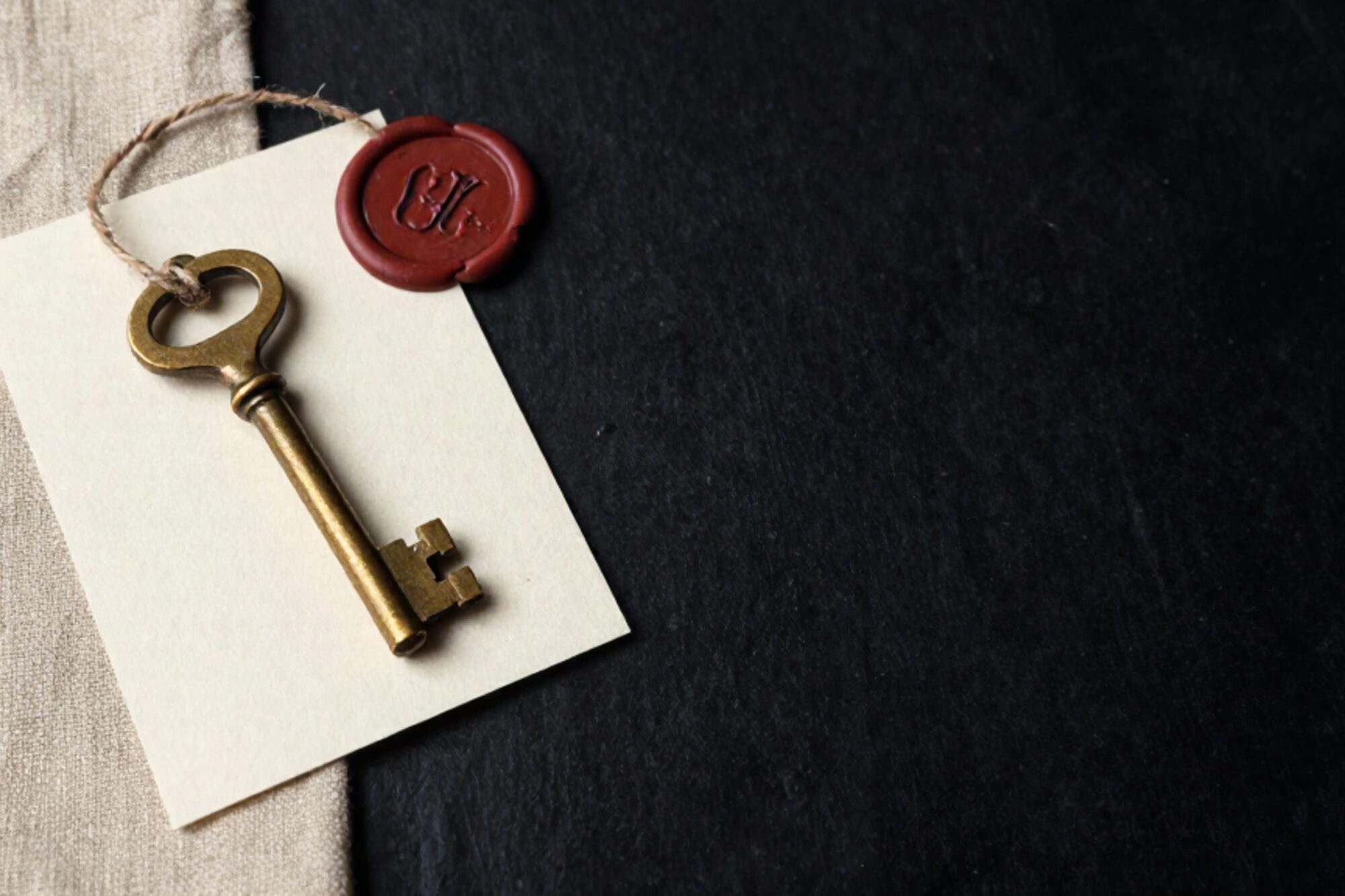 Brass key with twine loop on cream card stamped with serial number, red wax seal in corner, on bone linen.