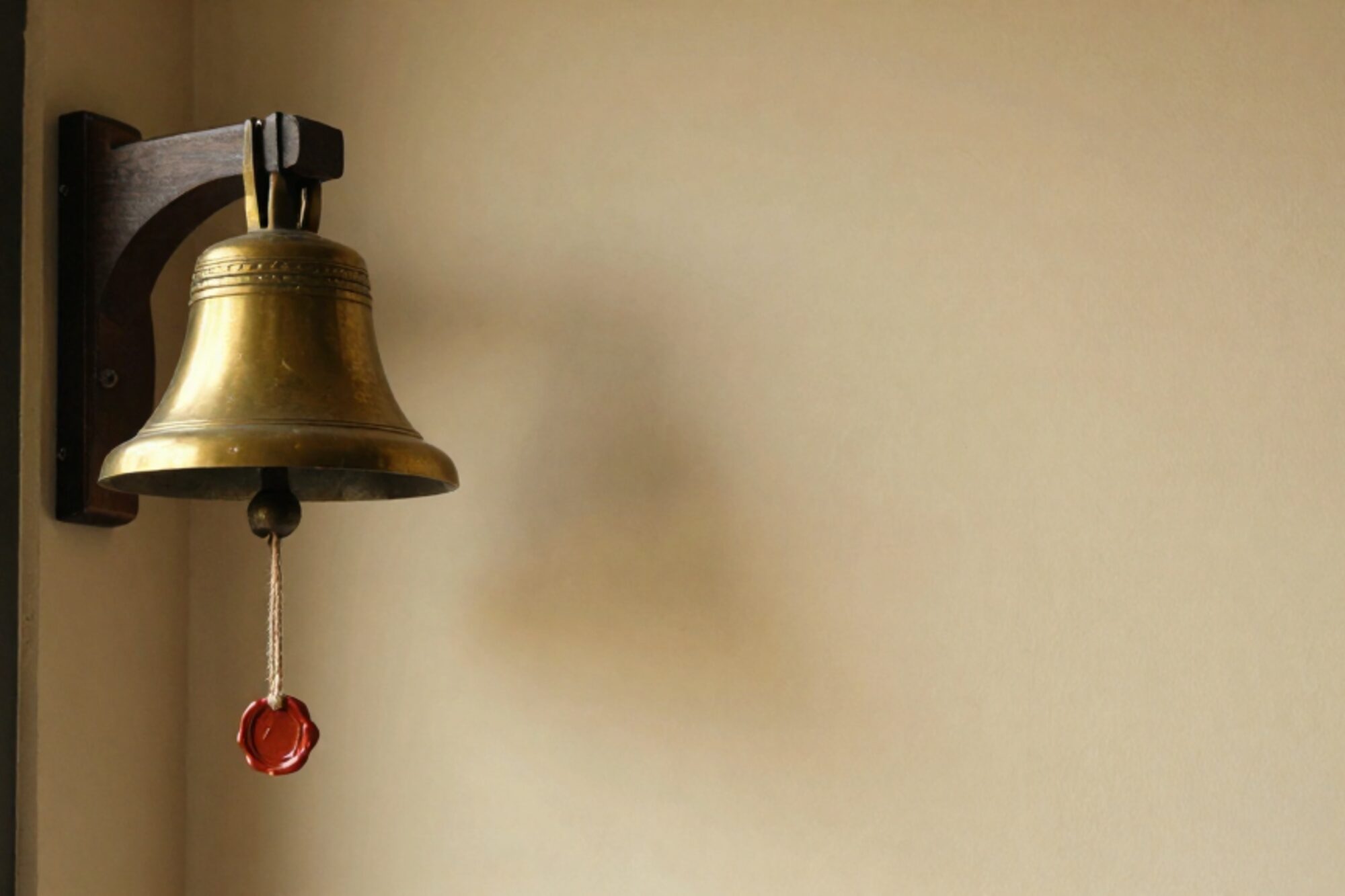 Brass ship's bell on dark oak bracket against bone plaster wall, hemp lanyard with clay-red wax-sealed paper tag.