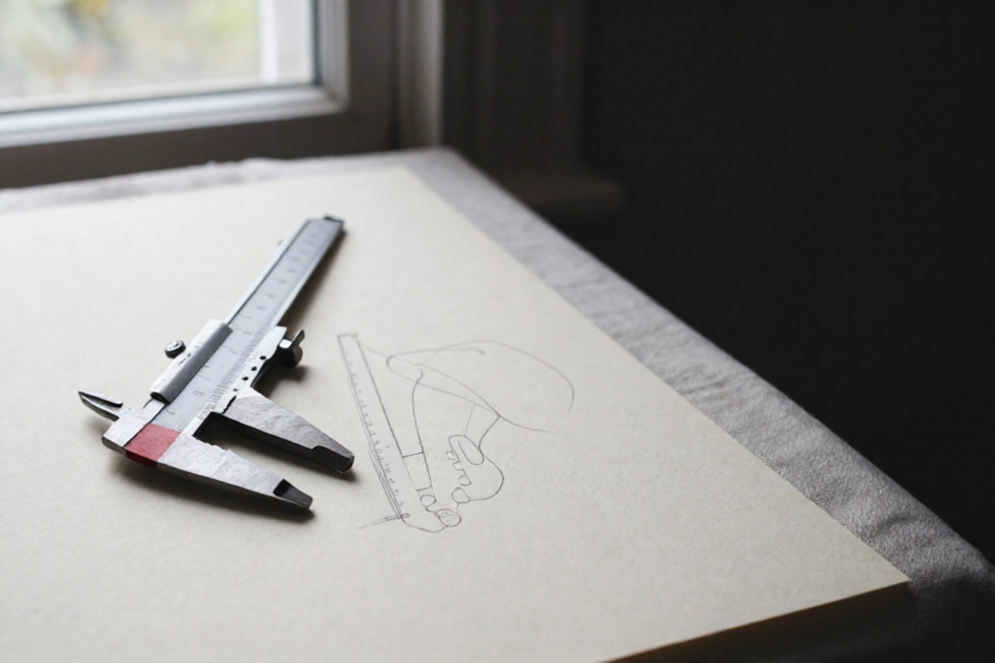 Machinist calipers on cream paper template with pencil lines, clay-red slip under one jaw, scarred oak table, side window light.