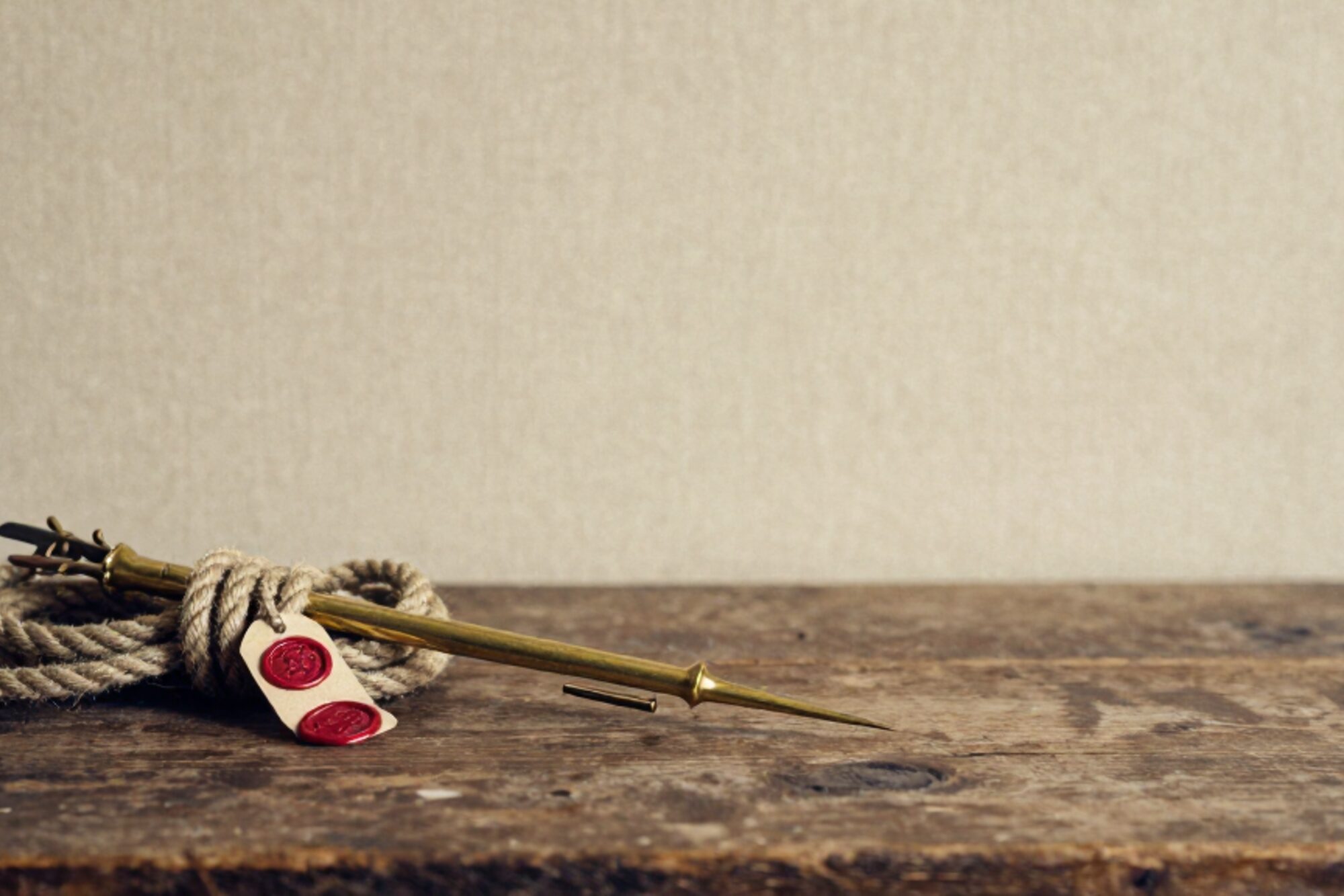 Brass marlin spike on coiled tarred hemp rope, strands picked apart, red wax seal on paper tag, oak plank, linen backdrop.